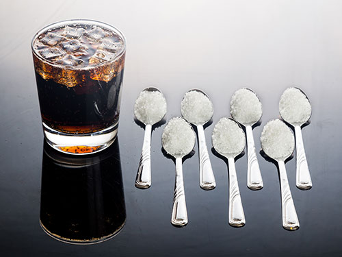 Photo: Seven teaspoons filled with sugar next to a glass of soda.