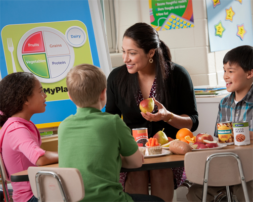 Photo: Teacher holding a fruit, and group of children sitting together at a table.