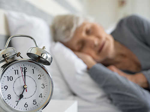 A clock in the foreground and in the background a senior female sleeping.